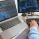 Side View Of Programmer Typing On Keyboard Looking At Many Monitors